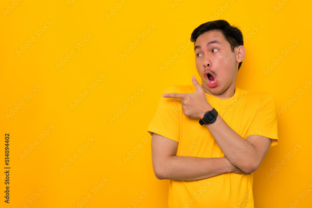 Shocked young Asian man in casual t-shirt pointing finger aside at copy space isolated on yellow background. Great offer concept