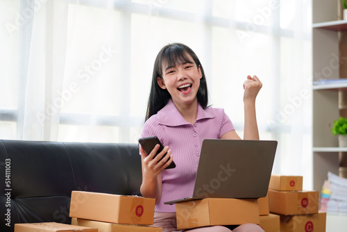 Young Asian small business owner successful selling online and holding smartphone with laptop computer. Happy Asian business woman successful excited raised hands rejoicing.