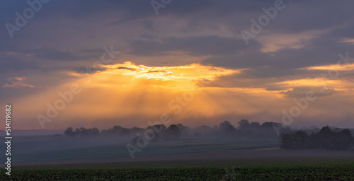 The first rays of the sun break through the fog and clouds and illuminate the green field