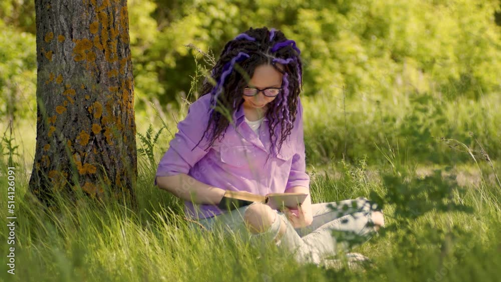 A woman in a purple shirt and glasses reads a literature under a tree. A girl is resting with a book in the forest. Female with a book is sitting on the grass on a clear summer day A woman in a purple shirt and glasses reads a literature under a tree. A girl is resting with a book in the forest. Female with a book is sitting on the grass on a clear summer day