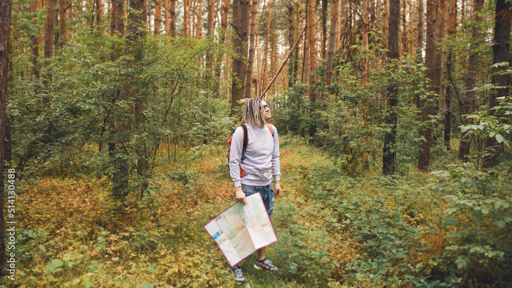 A traveling man with dreadlocks with a map in the forest. A male traveler with a backpack and a map in his hand in the forest in cloudy weather A traveling man with dreadlocks with a map in the forest. A male traveler with a backpack and a map in his hand in the forest in cloudy weather