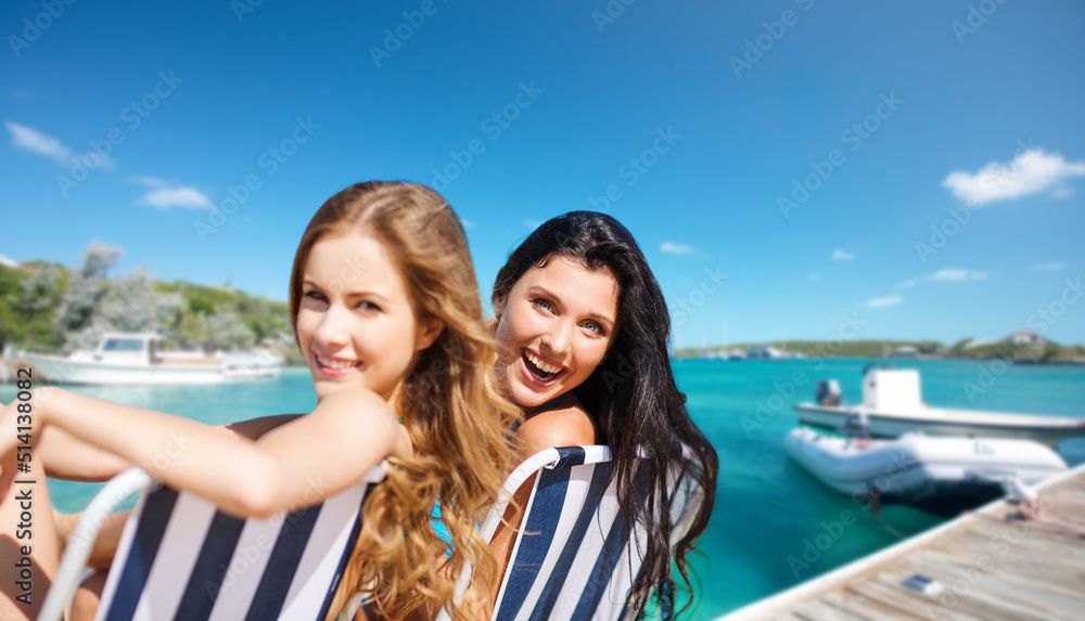 travel, tourism and summer vacation concept - beautiful happy women or female friends sunbathing on beach chairs over wooden pier and boat on tropical background in french polynesia
