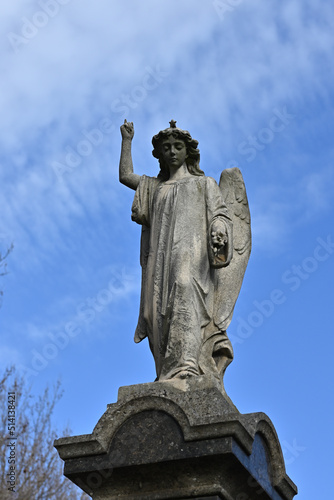 Worn stone sculpture of an angel pointing skyward while looking down, with blue sky and light clouds in the background