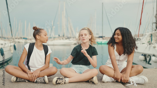 Laughing three girls friends pre-teenage sitting on the waterfront against ships and yachts background. Teenagers singing and clapping on the outdoors in seascape background © Andrii Nekrasov