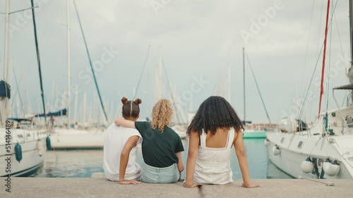 Laughing three girls friends pre-teenage sitting on the waterfront against ships and yachts background. Teenagers singing and clapping on the outdoors in seascape background