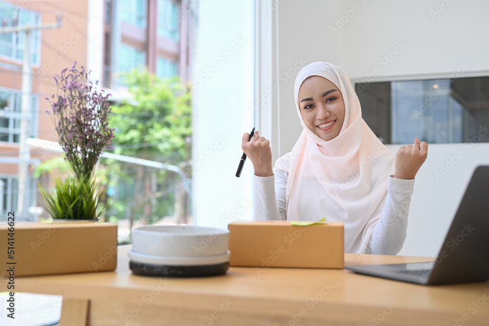 Happy young Muslim woman raising hands up, celebrating after finish big order from customer. Online selling, e-commerce Happy young Muslim woman raising hands up, celebrating after finish big order from customer. Online selling, e-commerce