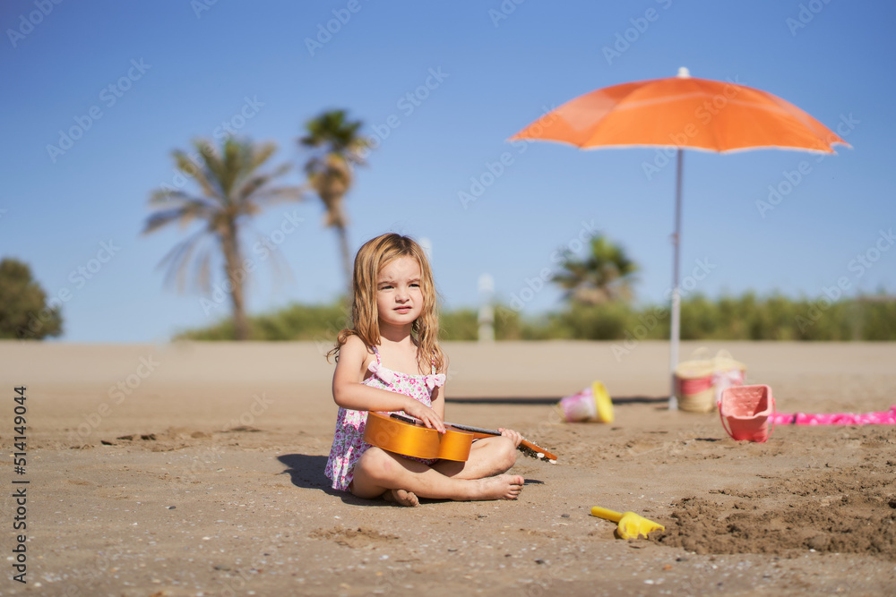 a little blonde girl playing a ukulele sitting on the sand on the beach