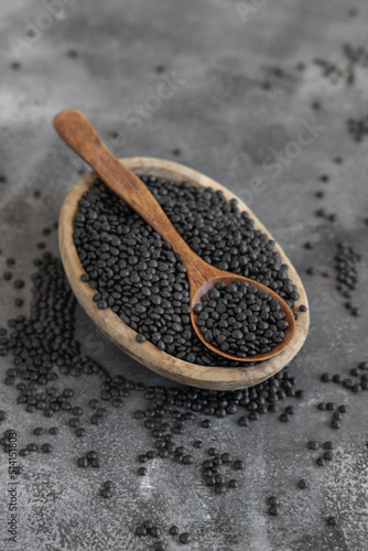 Bowl of dry black lentils beans with a spoon on grey table close up, protein source for vegetarian diet