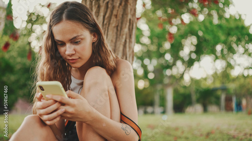 Beautiful girl with long wavy hair wearing in white top uses mobile phone while sitting on the green grass under tree in the park. Girl flip through the pictures on your phone with fingers