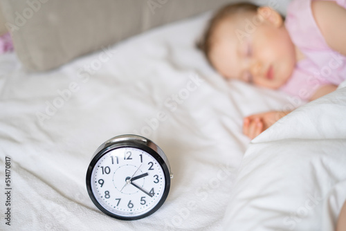 A little girl sleeps in a white bed with a clock. Sleep schedule for a child. Alarm feeding