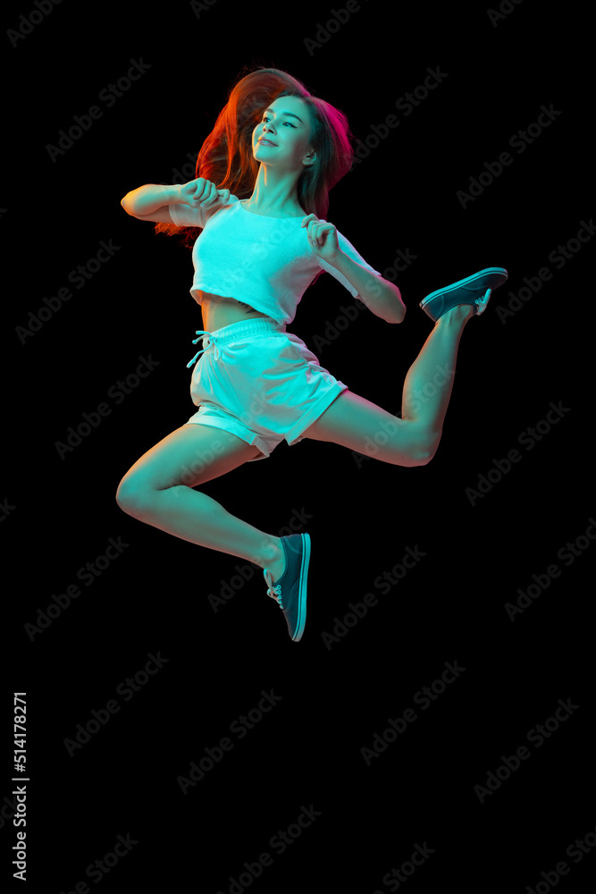 Full-length portrait of young cheerful girl posing, jumping isolated over black studio background in neon light