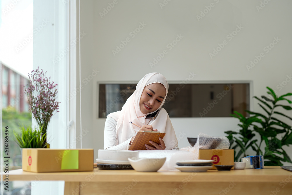 Young asian muslim woman business owner with parcel boxes online sales job, calling with smartphone, get an order from customers, take notes, and make arrangements for delivery by post. Young asian muslim woman business owner with parcel boxes online sales job, calling with smartphone, get an order from customers, take notes, and make arrangements for delivery by post.
