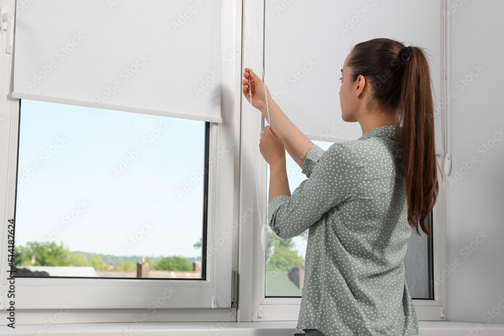 Woman opening white roller blind on window indoors
