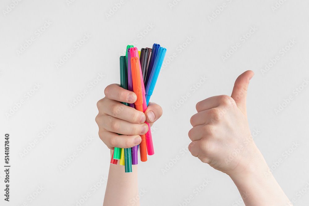 child hand holding many colorful felt-tip pens and showing thumbs up above a white and gray background, copy space - obrazy, fototapety, plakaty 