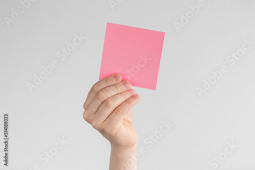 child hand holding a square pink blank reminder or paper notes above a white and gray background, copy space