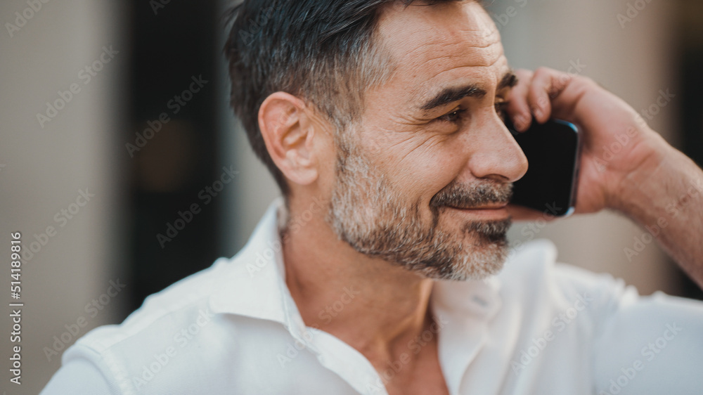 Obraz premium Close-up mature businessman with neat beard uses mobile phone sits on bench in the financial district in the city. Successful man talking on smartphone.