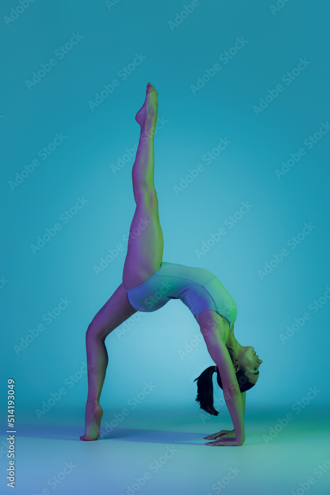 Portrait of young sportive girl doing stretching exercises isolated over blue studio background in neon light. Keeping strong, slim figure