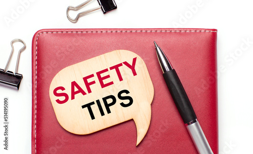 On a light background there are black paper clips, a pen, a burgundy notepad a wooden board with the text SAFETY TIPS