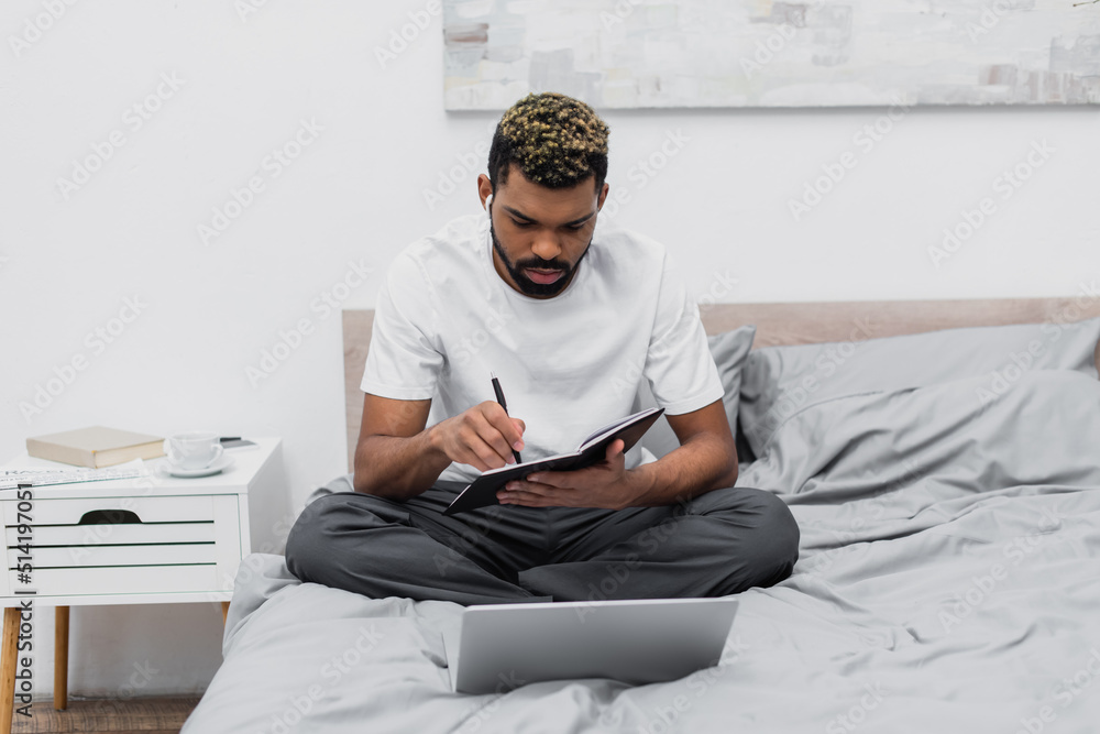bearded african american man in wireless earphones looking at notebook near laptop on bed.