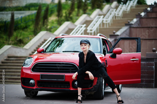 Portrait of beautiful blonde sexy fashion woman model in cap and in all black with bright makeup near red city car.