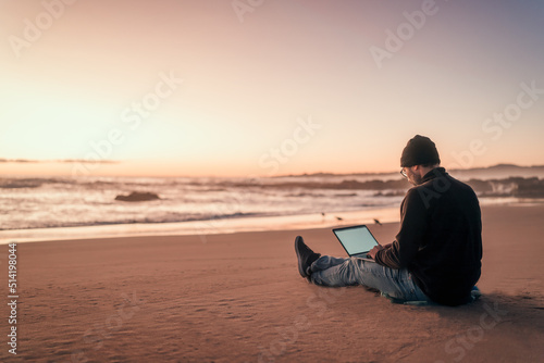 silhouette of a person working on his laptop outdoors on the beach at golden hour, back view. photo