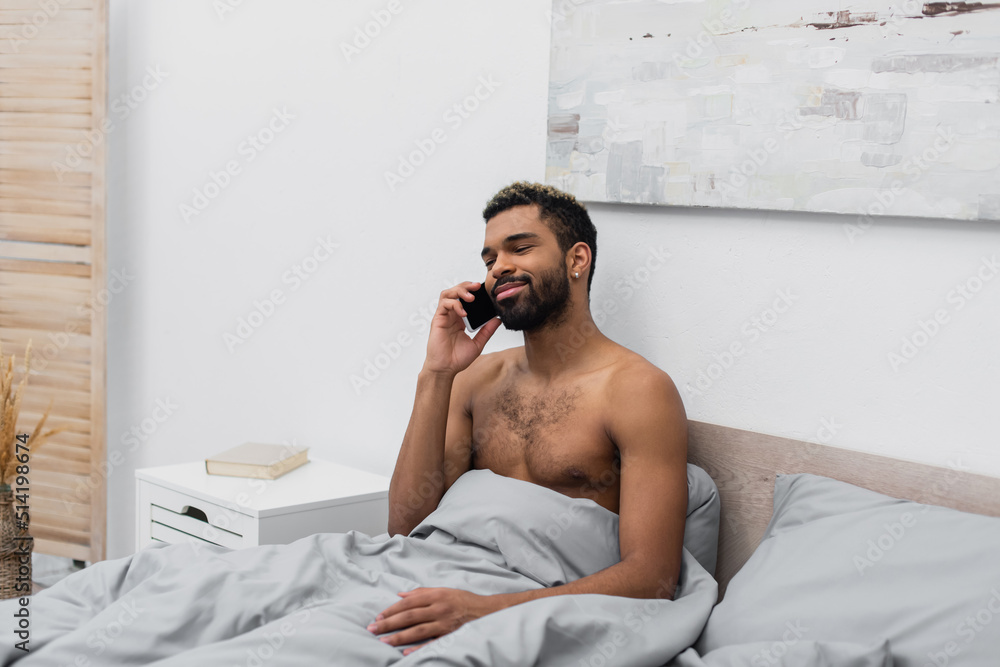 shirtless and happy african american man with dyed hair talking on mobile phone in bed. shirtless and happy african american man with dyed hair talking on mobile phone in bed.