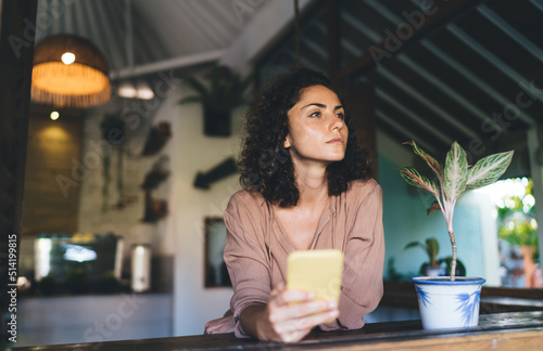 Pensive hipster girl with modern cellphone technology thoughtful looking away thinking about content planning during daytime at terrace, pondering female blogger with digital smartphone gadget