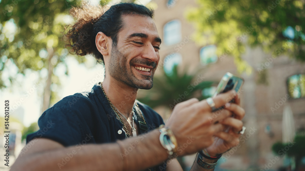 Attractive italian guy with ponytail and stubble is using mobile phone sitting on bench at old buildings background. Stylish man writes message, views photo on smartphone Attractive italian guy with ponytail and stubble is using mobile phone sitting on bench at old buildings background. Stylish man writes message, views photo on smartphone