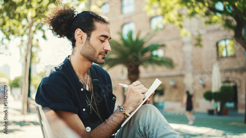 Young italian guy with ponytail and stubble sits on street bench and makes sketches with pen on piece of paper