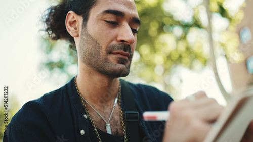 Young italian guy with ponytail and stubble sits on street bench and makes sketches with pen on piece of paper