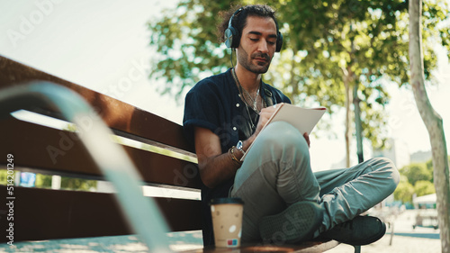 Young italian guy with ponytail and stubble sits with headphones on street bench listens to music and makes sketches with pen on piece of paper on cityscape background