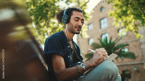Young italian guy with ponytail and stubble sits with headphones on street bench listens to music and makes sketches with pen on piece of paper on cityscape background