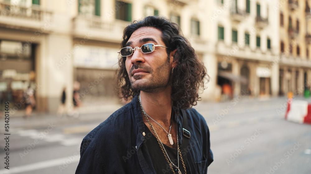 Young italian guy with long curly hair and stubble walks smiling down the street. Stylish man with sunglasses and lot of chains smiling and looking around Young italian guy with long curly hair and stubble walks smiling down the street. Stylish man with sunglasses and lot of chains smiling and looking around