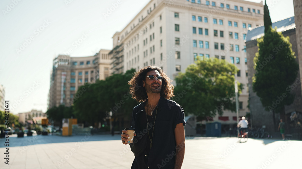 Young italian guy with long curly hair and stubble is enjoying aromatic coffee. Stylish man drinks coffee on old town background
