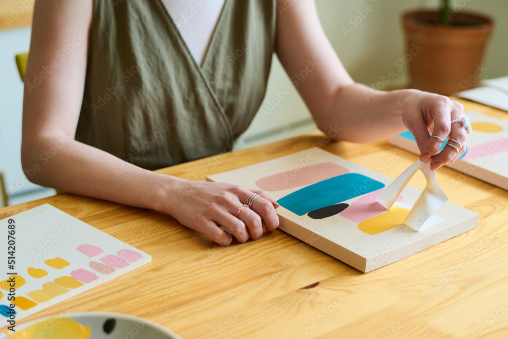 Hands of young creative female sitting by wooden table in workshop or studio and using stencil while creating new abstract painting Hands of young creative female sitting by wooden table in workshop or studio and using stencil while creating new abstract painting