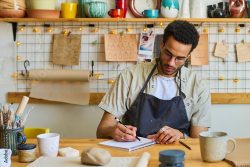 Young creative black man drawing sketches of new handmade craft items in notepad or making working notes while sitting by table in studio photo