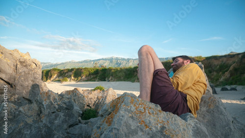 Hombre blanco tumbado en una roca de playa con montañas en el horizonte