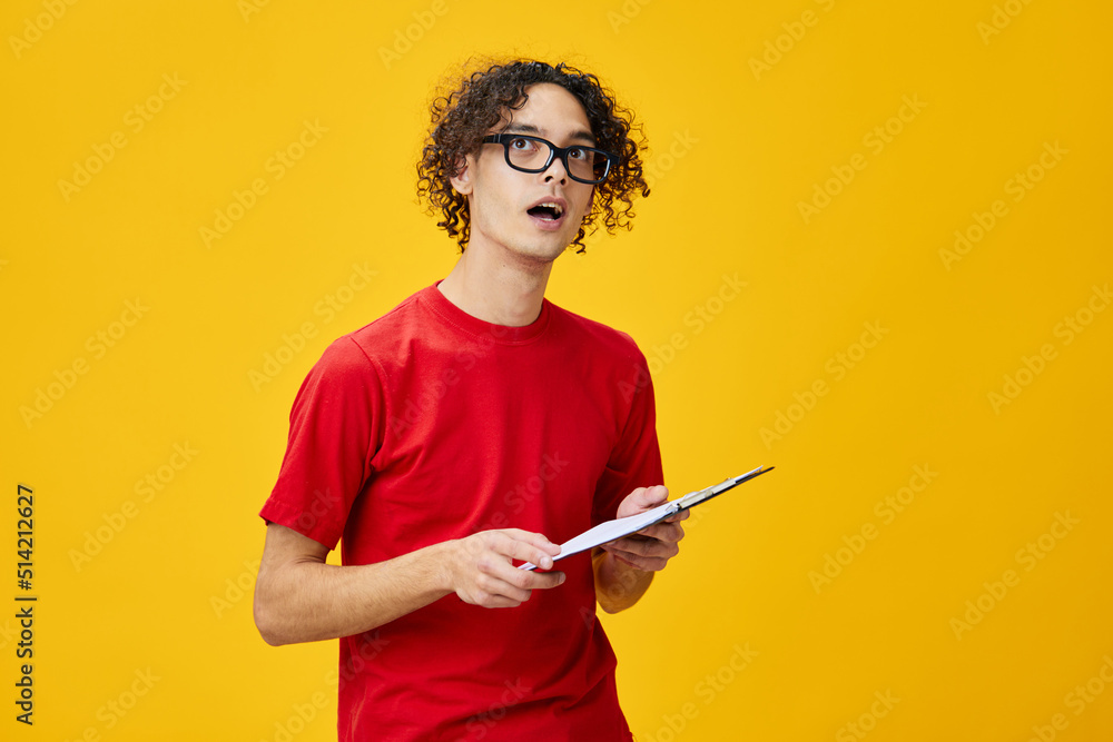 Confused myopic young student man in red t-shirt funny eyewear holds tablet folder with study notes looks aside posing isolated on over yellow background. Free place for ad. Education College concept Confused myopic young student man in red t-shirt funny eyewear holds tablet folder with study notes looks aside posing isolated on over yellow background. Free place for ad. Education College concept