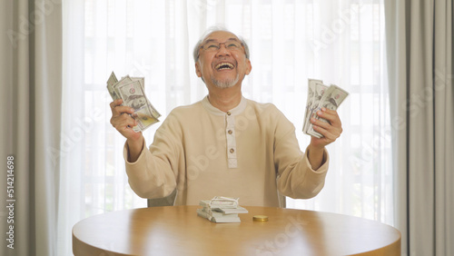 Portrait of happy rich Asian Thai old senior retired man counting money to save banknote in financial account and investment concept on table at home or house. People lifestyle. Win lottery