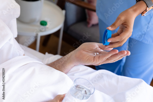 Mid section of biracial female health worker giving medical pills to caucasian senior man on the bed