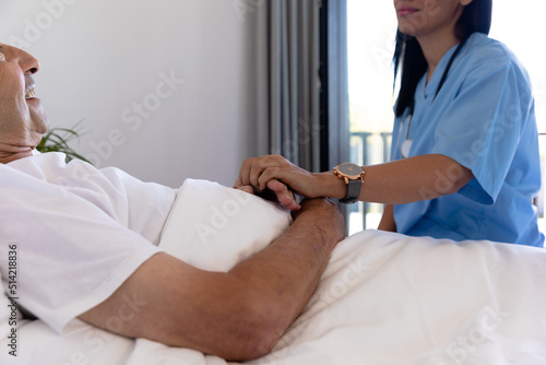 Mid section of biracial female health worker supporting caucasian senior man lying on the bed