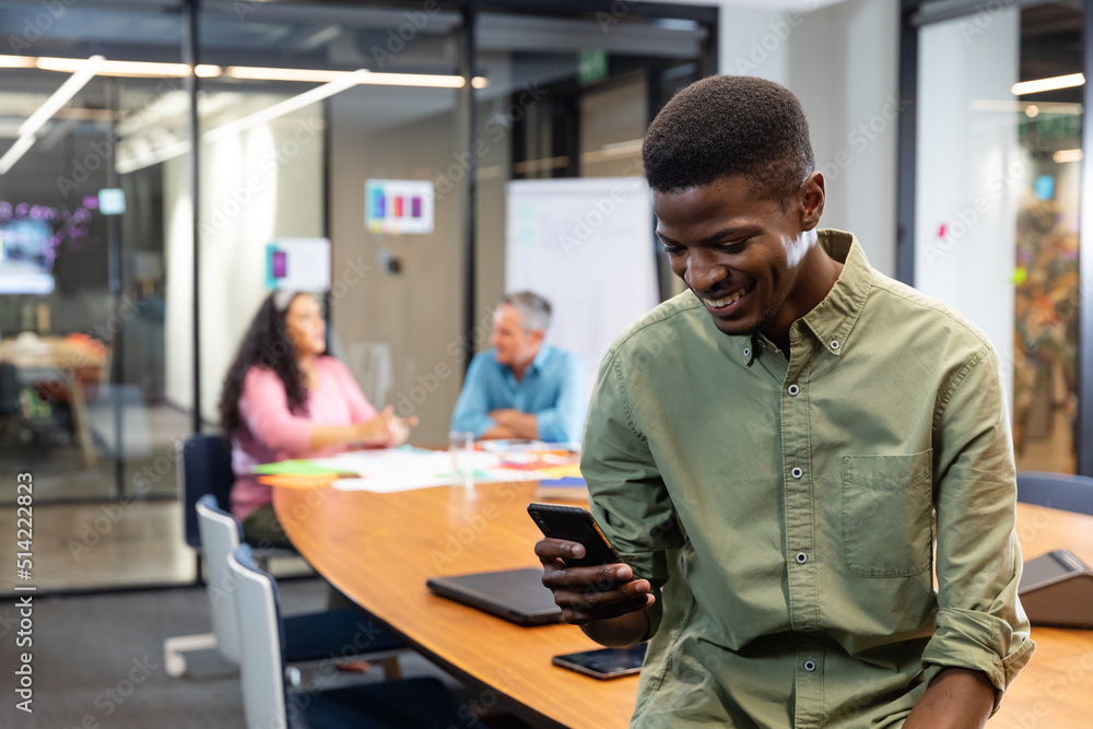 Smiling young african american businessman using smart phone while colleagues in background Smiling young african american businessman using smart phone while colleagues in background
