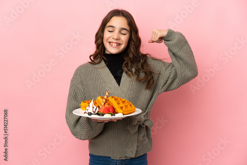 Little girl holding waffles isolated on pink background doing strong gesture