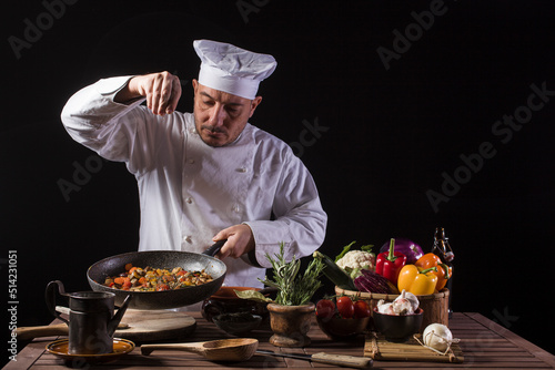 Male chef in white uniform and hat putting salt and herbs on food plate with vegetable