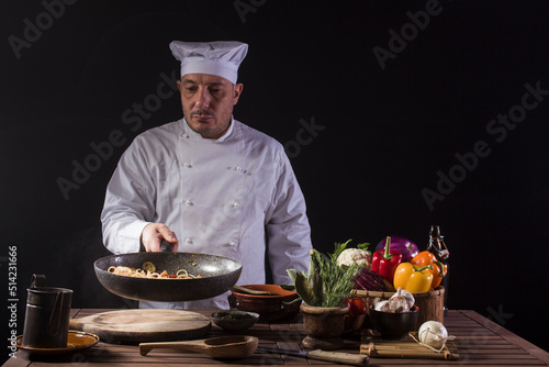 Male chef in white uniform holding a frying pan, sautéing spaghetti with fresh vegetables flying in the air