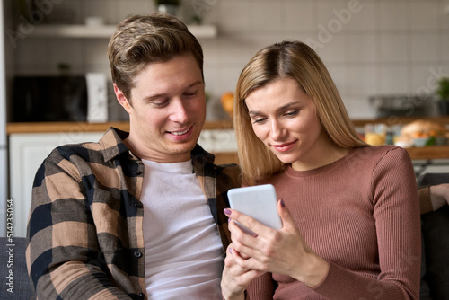 Young happy couple holding smartphone using mobile cell phone apps at home, doing ecommerce online shopping together, ordering food, checking social media or reading news in chats together.