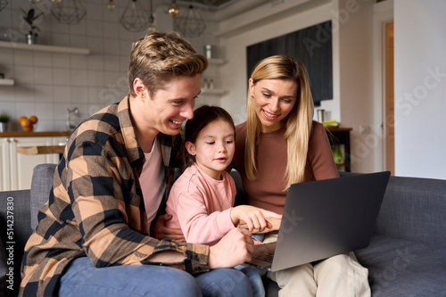 Happy family young parents and kid child daughter using laptop computer technology device having fun sitting on couch at home, talking, laughing doing ecommerce shopping online, e learning together.