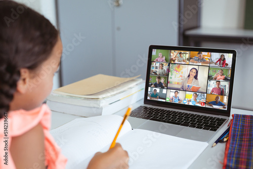 Asian girl with book and pencil attending online class over laptop on table at home