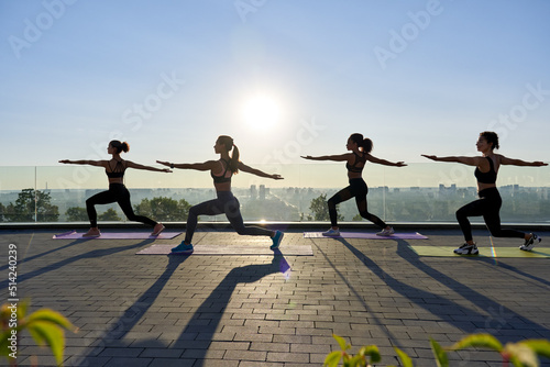 Female silhouettes stand in yoga virabhadrasana warrior pose at group class outdoor on sunrise. Multiethnic fit healthy young women do yoga fitness exercise together at retreat with sunset summer sky.