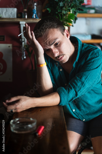 Portrait of young man looking worried smoking cigarette at pub or bar. Nicotine addicted person with bad habits, sitting lonely and thoughtful, leaning head on his hand